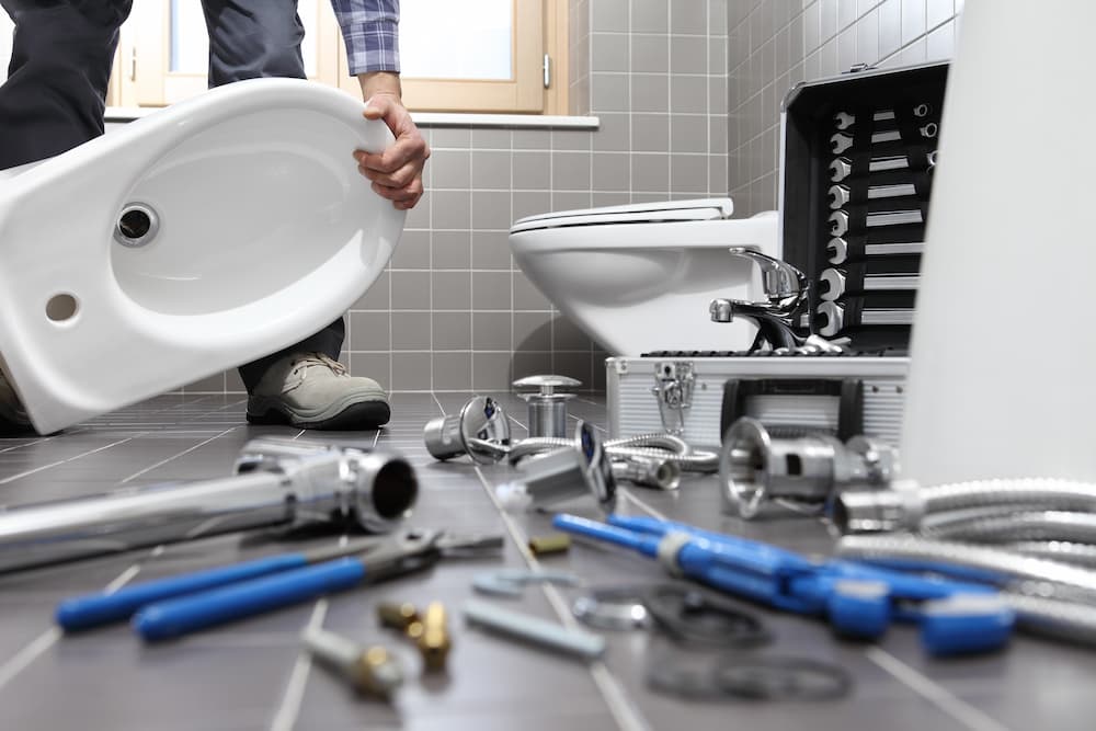 Plumber installing a new white bidet or sink basin in a modern grey bathroom with tools scattered on the floor. 
